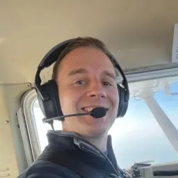 Joe Charman in the cockpit of a light aircraft, wearing a headset, with the wing strut and sky through the window behind.